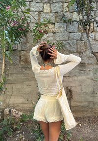 Bela, kvačkana jopica z zvonastimi rokavi, združena z belimi hlačami z čipkastim robom. Bež toaletna torba visi z rame.