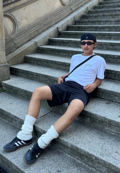 Conjunto casual que incluye una camiseta blanca, pantalones cortos negros, zapatillas negras con detalles blancos y calcetines blancos hasta la rodilla, sentado en unos escalones de piedra.