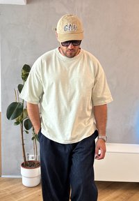 Light green oversized t-shirt, relaxed fit, textured fabric; dark blue wide-leg pants; beige cap with "GAP" logo; dark sunglasses.