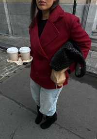 Manteau rouge en laine à double boutonnage, sac noir tissé, jeans bleu clair, bottes noires montantes, tenant deux gobelets en papier dans un porte-gobelets beige.