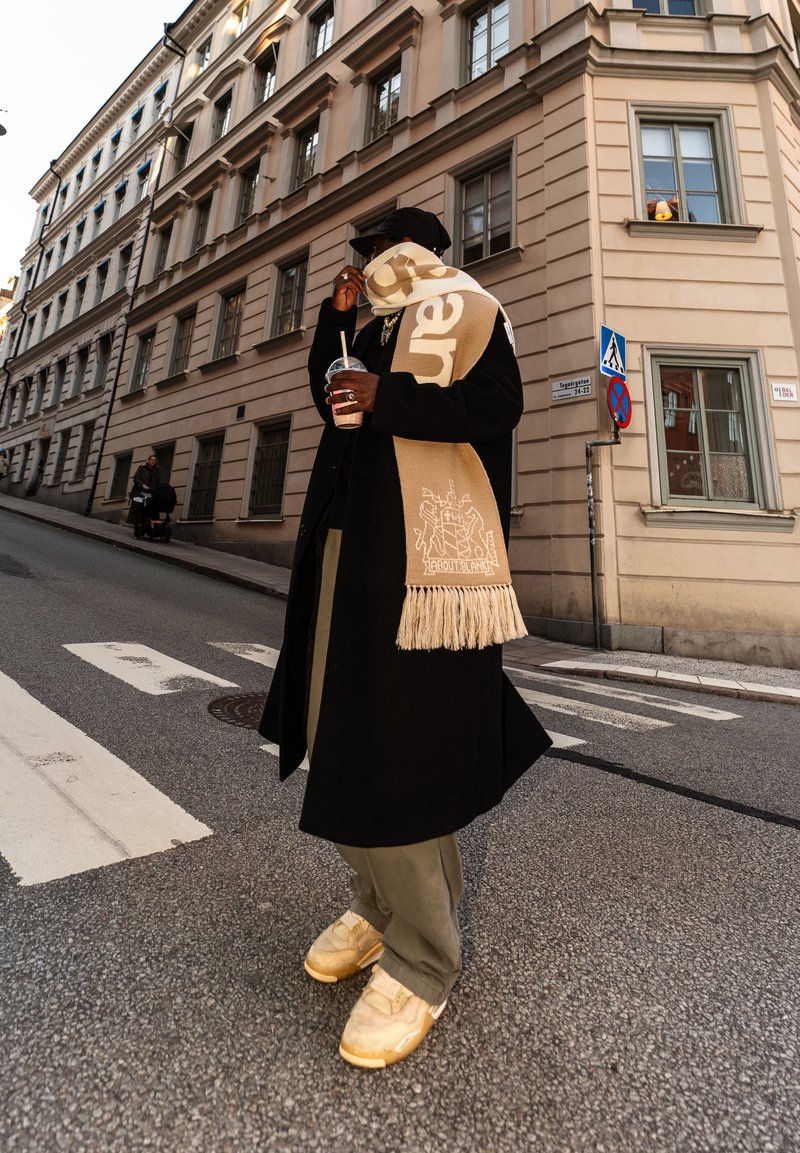 Personne en manteau noir et casquette avec une écharpe beige, tenant une boisson, debout dans une rue de la ville près d'un passage pour piétons et d'un bâtiment historique.