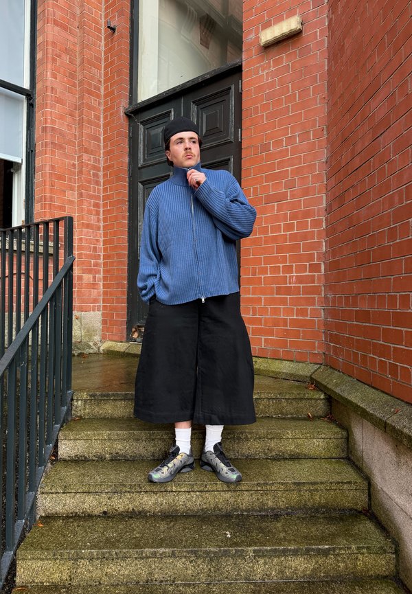 Jeune homme portant un pull à fermeture éclair bleu, pantalon large noir, chaussettes blanches, baskets et une casquette noire, debout sur des marches en pierre près d'un bâtiment en briques rouges.