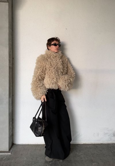Persona con chaqueta de piel rizada beige, pantalones anchos negros, gafas de sol negras, sosteniendo un bolso negro, de pie frente a una pared blanca texturizada.