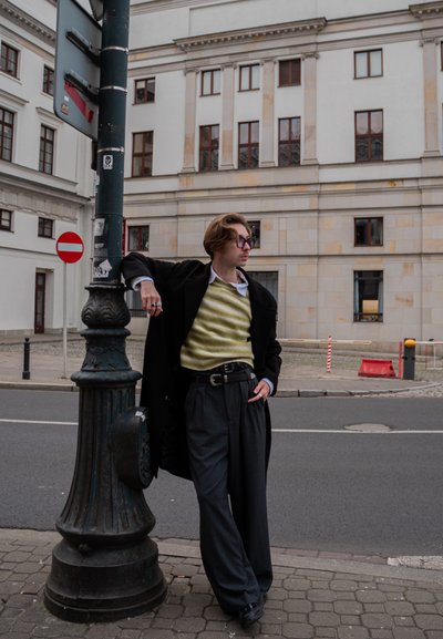 Abrigo negro, suéter verde a rayas, camisa blanca y pantalones anchos grises. De pie junto a una farola con arquitectura moderna cercana.