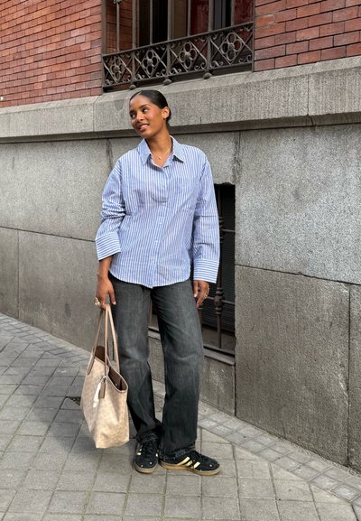 Camisa de botones a rayas azul y blanca, pantalones grises, zapatillas negras con acentos amarillos y una bolsa de mano ligera de color beige. Texturas suaves y ajuste relajado.