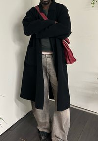 Άτομο στέκεται σε εσωτερικό χώρο φορώντας μακρύ μαύρο παλτό, γκρι τζιν, μαύρα παπούτσια και κρατώντας μια κόκκινη τσάντα ώμου με σταυρωμένα τα χέρια.