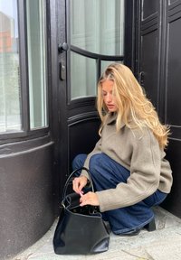 Fekete bőr kézitáska lekerekített fogantyúkkal, egy szürke kőre helyezve. Az illető világosbarna fleecdzsekit és kék farmert visel, és egy ajtó mellett guggol.