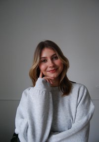 Helle graue Sweatshirt mit übergroßen Ärmeln. Die Trägerin hat schulterlanges, welliges braunes Haar und berührt mit ihrer rechten Hand ihre Wange.