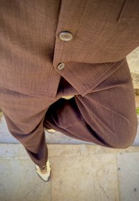 Sacou dintr-un material texturat, de culoare maro, cu două butoane și pantaloni asortați. Pantofii negri și crem completează ținuta.