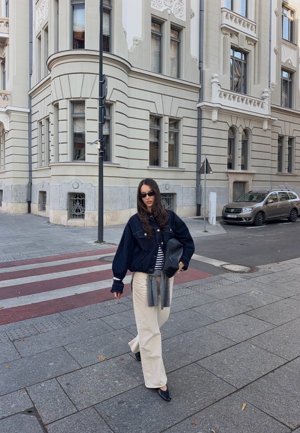 Une femme portant des lunettes de soleil, une veste sombre et un pantalon crème marche dans une rue de la ville, à côté d’un bâtiment orné en pierre claire.