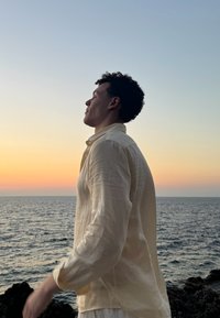 Ανοιχτό μπεζ λινό πουκάμισο με χαλαρή εφαρμογή, με διπλωμένα μανίκια, φορεμένο από ένα άτομο κοντά στη θάλασσα κατά τη διάρκεια του ηλιοβασιλέματος, με ήρεμη ατμόσφαιρα.