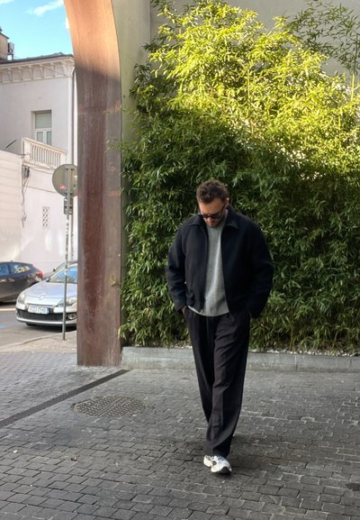 Chaqueta negra, suéter gris y pantalones oscuros; caminando sobre adoquines con vegetación al fondo. Zapatillas deportivas casuales en blanco y gris.
