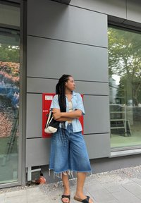 Kék denim culottok, rojtos szegéllyel, világoskék csíkos, rövid ujjú ing, fekete papucs és fekete-fehér válltáska. Piros tűzjelző doboz a háttérben.