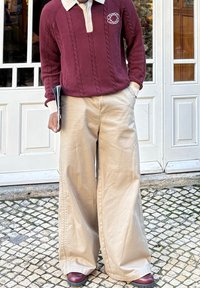 Personne portant un pull torsadé bordeaux et un pantalon large beige, debout sur des pavés devant des portes blanches à panneaux, tenant un magazine plié.