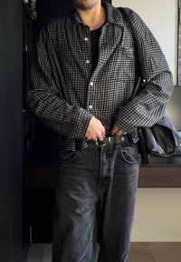 Homme portant une chemise à carreaux noire et grise, un jean noir, ajustant une ceinture noire, portant un grand sac à bandoulière en cuir noir à l'intérieur.