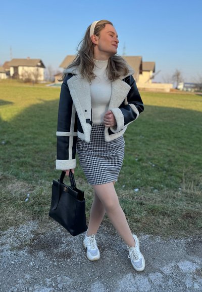 Falda de pata de gallo en blanco y negro, suéter de cuello alto crema, chaqueta de cuero negro y piel de oveja, bolso negro y zapatillas blancas con cordones. Fondo de hierba y cielo.