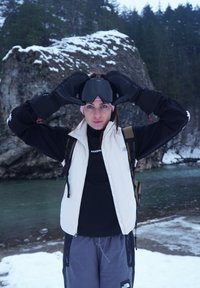 Femme en tenue d'hiver tenant des lunettes de ski au-dessus de ses yeux, debout près d'une rivière enneigée et d'un paysage rocheux avec des arbres.