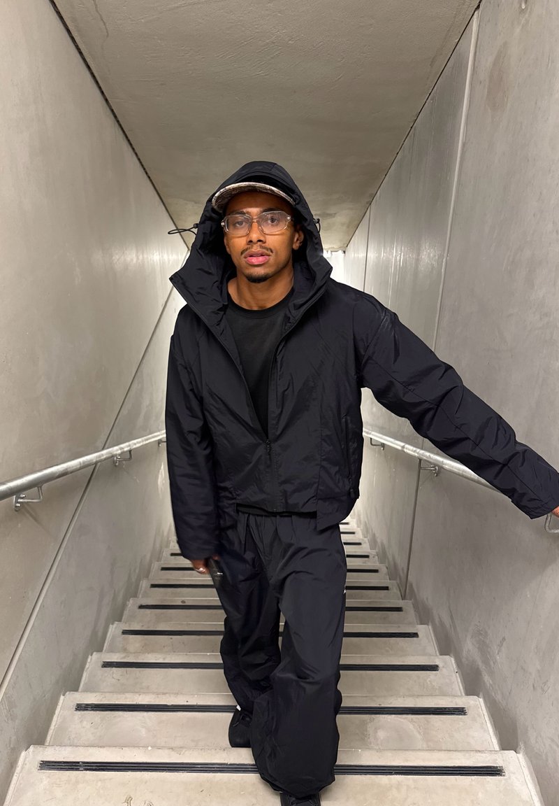 Veste et pantalon noirs, imperméables et à coupe décontractée. Lunettes transparentes, casquette à motifs, et un escalier avec rampe en métal dans un couloir gris.