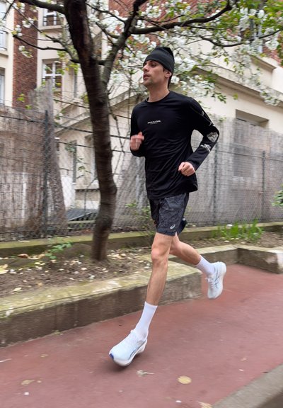 Hombre con ropa deportiva negra y zapatos blancos corriendo en un camino junto a árboles y una valla de alambre en una zona urbana.