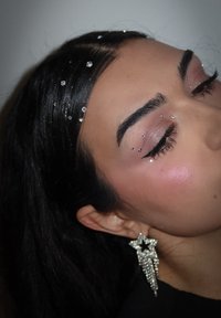Close-up of a face with dark hair adorned with rhinestones, featuring shimmering eye makeup and a silver star-shaped earring with fringe.