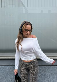 Fehér hosszú ujjú, off-shoulder felső, állatmintás szűk farmer, fekete fejhallgató és fekete táska; egyszerű póz tompa háttér előtt.