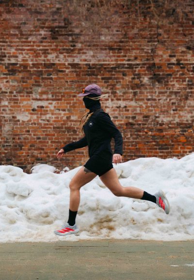 Persona con ropa de correr negra, gorra morada y mascarilla trotando por un camino nevado junto a un muro de ladrillos.