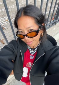 Svart gliderekke hettegenser over en rød NFL-drakt med hvite og gull aksenter, lagvis med en sølvkjedehalskjede og mørke, store solbriller.