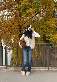 Modell iført en kremfarget strukturert vest over en hvit topp, mørkeblå jeans, lyse beige tøfler, og holder en brun veske, foran høstløvverk.