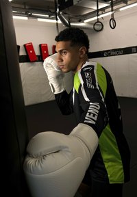 Bokseris, vilkintis baltus pirštines, stovi šalia juodo smūginio maišo. Marškiniai yra juodi su žaliais akcentais ir logotipu ant rankovės.