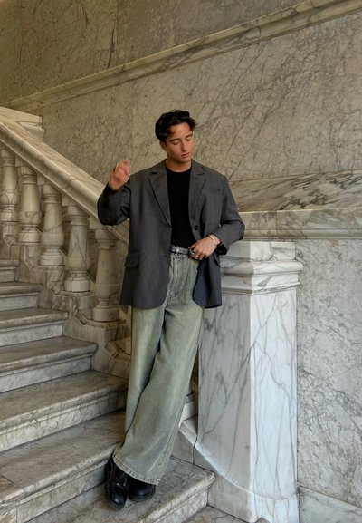 Blazer gris extragrande sobre camiseta negra, combinado con jeans de pierna ancha verdes claros y zapatos negros, posando frente a una escalera de mármol.