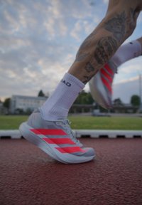 Gamba tatuata di un corridore con calzini bianchi e scarpe grigie con strisce rosa, catturata a metà passo su una pista rossa sotto un cielo nuvoloso.