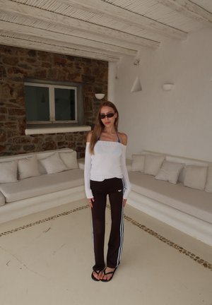 Mujer con gafas de sol, top blanco sin hombros, pantalones deportivos negros y chanclas, de pie en una habitación blanca con asientos acolchados y una pared de piedra.