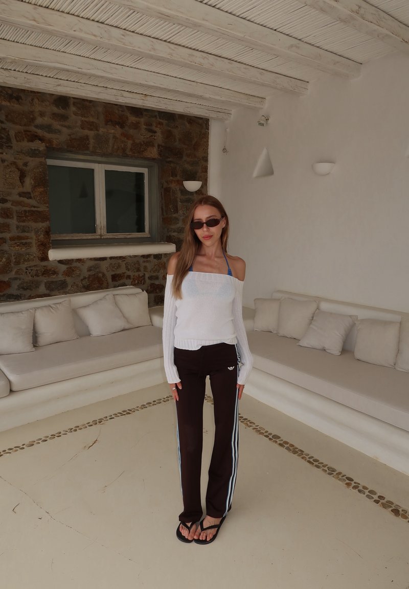 Femme portant des lunettes de soleil, un haut blanc à épaules dénudées, un pantalon de jogging noir et des tongs se tient dans une pièce blanche avec des sièges rembourrés et un mur en pierre.