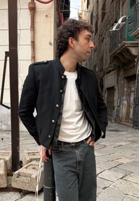 Veste militaire noire avec boutons argentés, portée sur un t-shirt blanc. Associée à un jean en denim foncé, debout devant un décor urbain texturé.