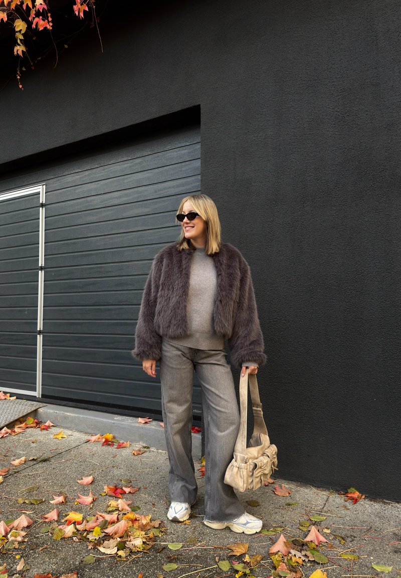 Mujer con gafas de sol, una chaqueta violeta esponjosa, pantalones grises y zapatillas blancas, sosteniendo un bolso beige, de pie en una acera cubierta de hojas.