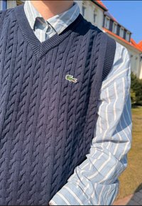 Temno modra pletena brezrokavnika z vzorcem kabla, z V-izrezom in zelenim logotipom krokodila Lacoste. Nošen preko svetlo modre črtaste srajce.