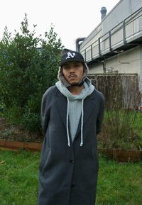 Cappotto di lana grigio con colletto a rever, sovrapposto a un maglione di maglia grigio chiaro con cappuccio. Berretto nero con logo e paraorecchie foderati in pelliccia.