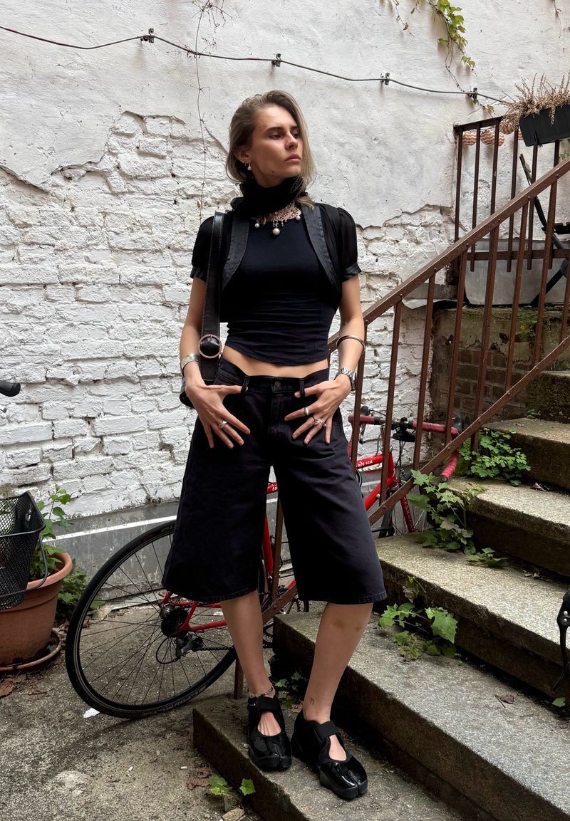 Femme vêtue tout en noir, debout les mains sur les hanches à côté d’un vélo rouge appuyé contre un mur en briques blanches et des escaliers en pierre avec du verdure.