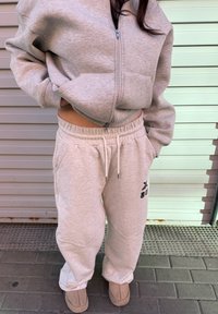 Personne portant un sweat à capuche gris clair zippé et un pantalon de survêtement assorti avec un texte noir sur la jambe gauche, debout sur le trottoir près d'un volet métallique.