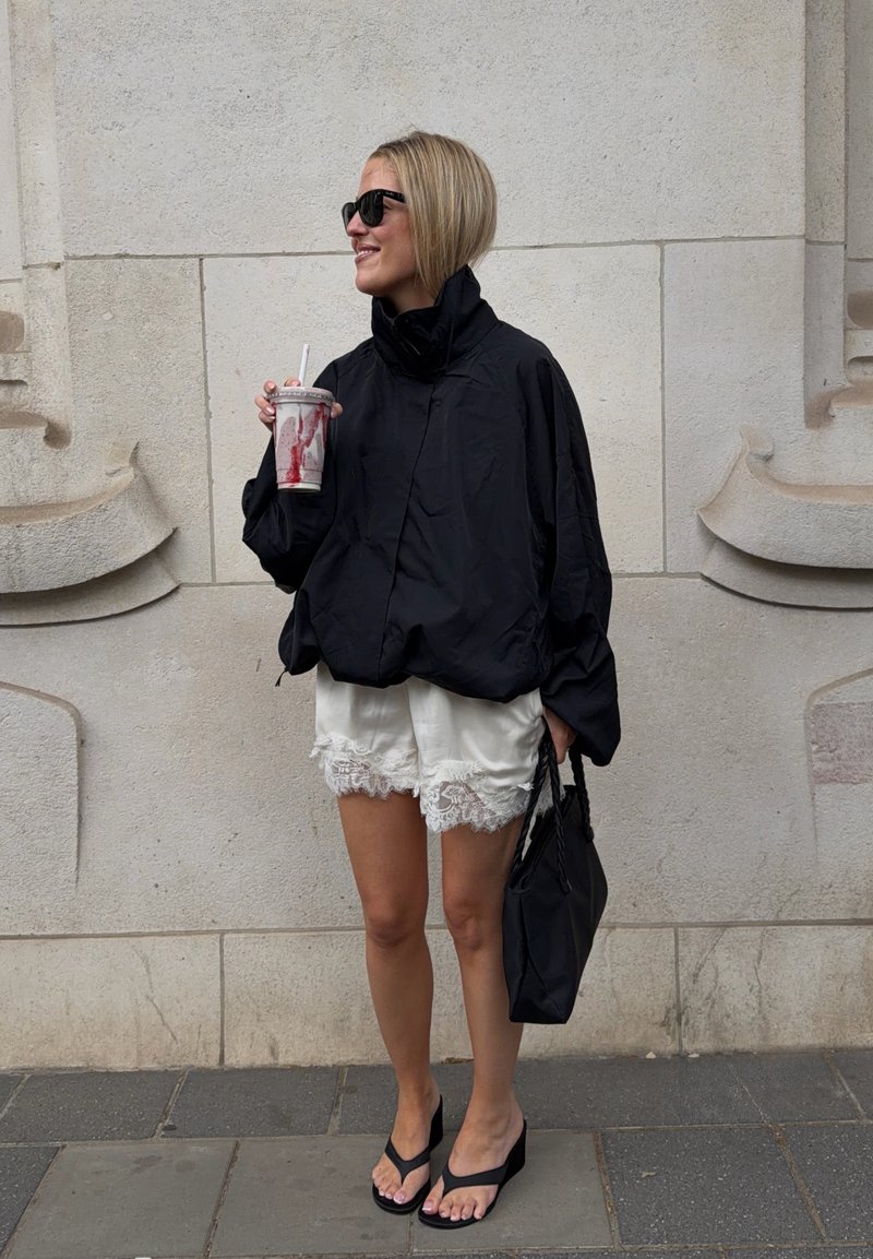 Femme debout près d'un mur en pierre avec des lunettes de soleil, une veste noire, un short en dentelle blanche, des sandales noires, tenant une boisson et un sac à main noir.