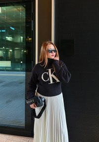 Femme en pull à col roulé noir et jupe plissée blanche tenant un sac noir, debout devant l'entrée d'un bâtiment moderne.