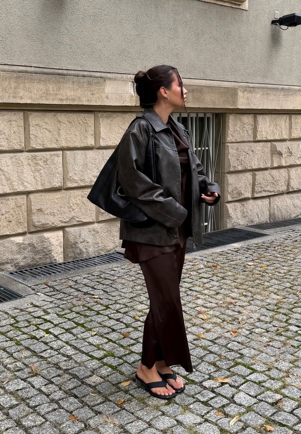 Mujer de pie en una calle empedrada con chaqueta de cuero negra, conjunto marrón, sandalias negras y llevando un bolso grande negro, cerca de un muro de piedra.