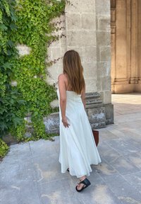 Weißes, ärmelloses Maxikleid mit einem fließenden Schnitt und dezenten Nähten. Getragen mit schwarzen Sandalen. Der Hintergrund zeigt eine Steinmauer und grüne Ranken.