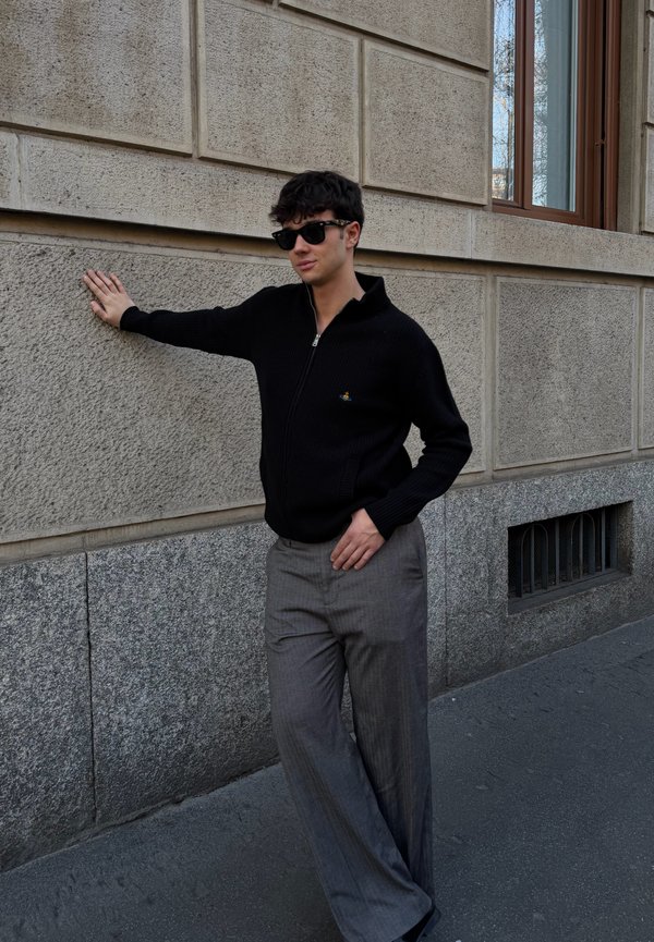 Ung mann med solbriller, svart zip-up genser og grå bukser som lener seg med én hånd mot en teksturert beige steinvegg på en bygate.