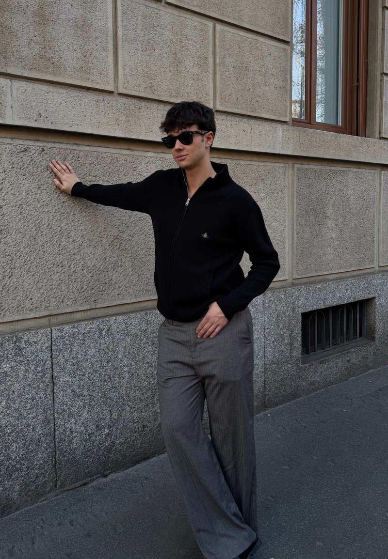 Jeune homme portant des lunettes de soleil, un pull noir à fermeture éclair et un pantalon gris, appuyé d'une main contre un mur en pierre beige texturé dans une rue de la ville.