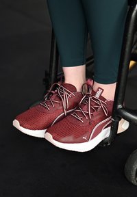 Wijnrode sportschoenen met een gestructureerd bovenwerk en witte accenten, voorzien van veters en merkopdruk, geplaatst naast een rolstoel.