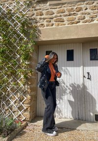 Zwarte leren motorjack, oranje brei-top, zwarte flared jeans en grijze sneakers. Achtergrond met een stenen muur en houten deuren.