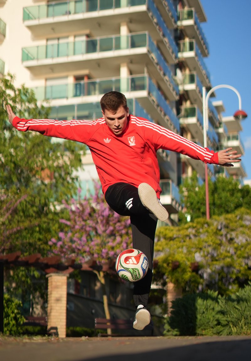 Jeune homme en veste rouge donnant un coup de pied à un ballon de football en plein air avec des bâtiments urbains et de la verdure en arrière-plan par une journée ensoleillée.