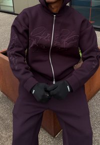 Sweat à capuche zippé violet foncé avec un motif en relief, présentant une coupe décontractée, des poignets côtelés, et accompagné d'un pantalon assorti et de gants noirs.