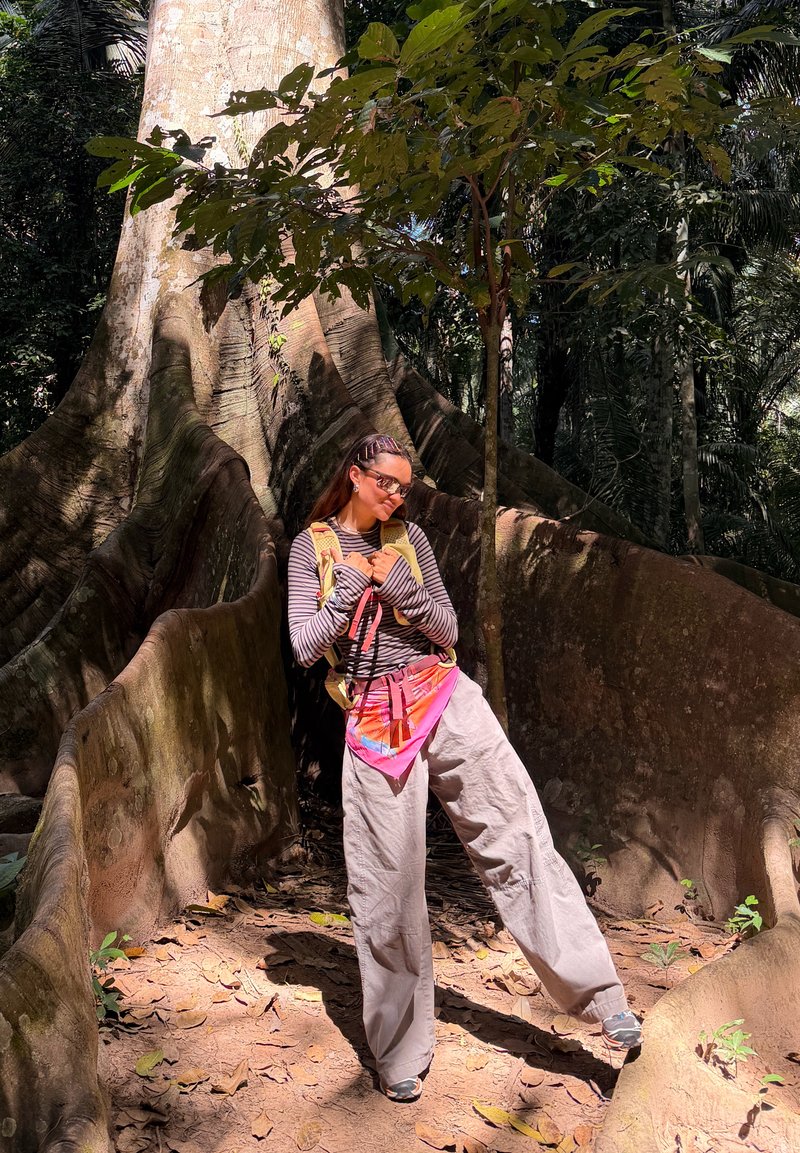 Femme en chemise rayée et pantalon beige debout parmi de grandes racines d'arbre dans une forêt ensoleillée, portant des lunettes de soleil et une sacoche colorée.